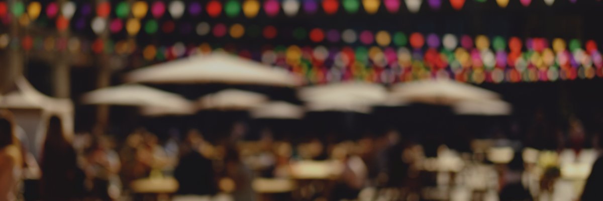 Mitmach-Angebote Bild 1 Unscharfe Aufnahme einer abendlichen Veranstaltung im Freien: Im Hintergrund hängen bunte Lichterketten, davor stehen mehrere helle Sonnenschirme. Darunter sind zahlreiche Menschen an Tischen zu erkennen, die eine lebendige Atmosphäre andeuten.